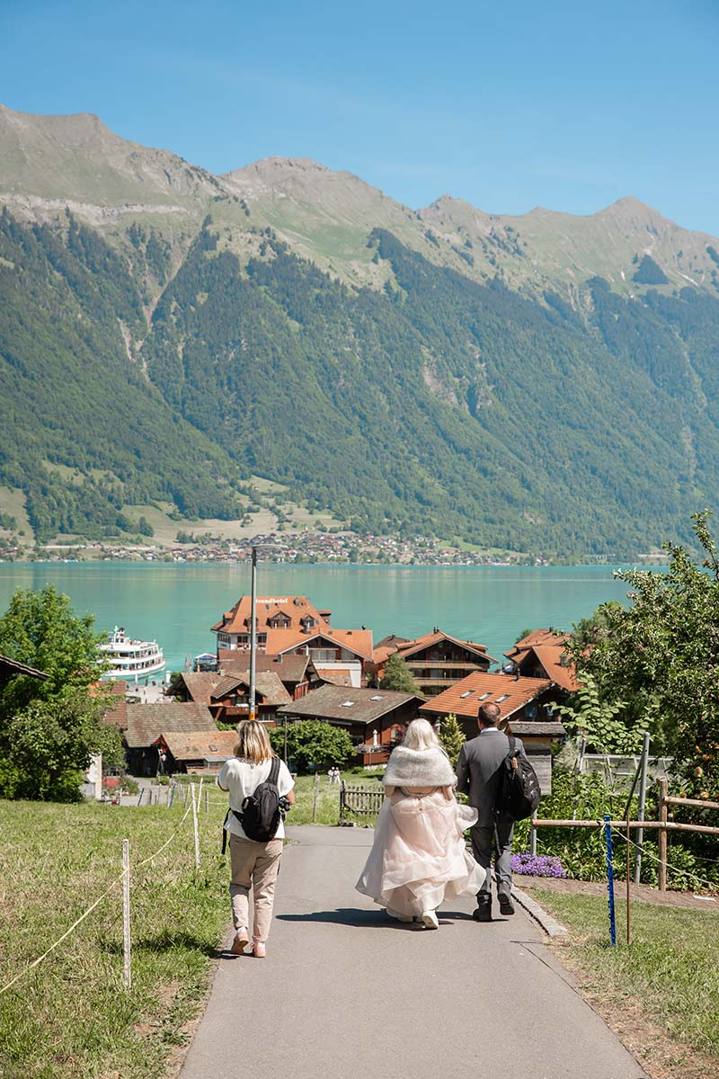 Three people walk down a paved path toward a Swiss lake, with a village and mountains in the background under a clear blue sky - perfect inspiration for your elopement page.