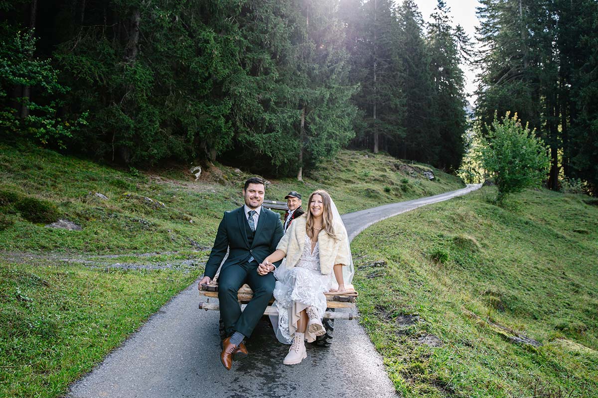 A bride and groom sit on a rustic wooden cart being pulled along by a tractor on a forest path in Wengen, surrounded by greenery and tall trees - an intimate scene perfect for an elopement