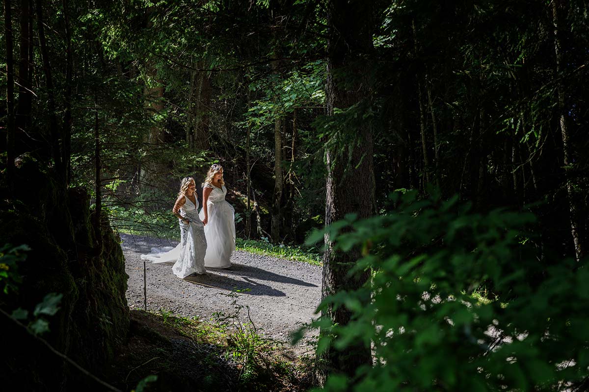 Two women in white dresses walk along a sunlit forest path, surrounded by tall trees and greenery—an intimate moment perfect for elopement inspiration or capturing memories from thoughtful elopement planning.