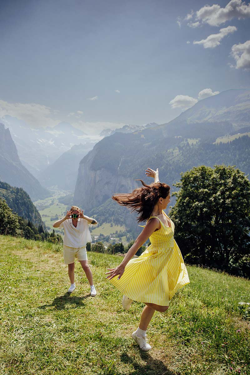 Two people are outdoors on a grassy hillside with mountains in the background; it’s a dreamy elopement moment - one in a yellow dress spinning joyfully, while the other stands nearby, holding a hat.