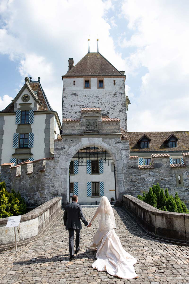 A bride and groom walk hand in hand toward the entrance of a historic stone building with blue-and-white shutters on a sunny day, capturing the romance and intimacy perfect for any elopement.