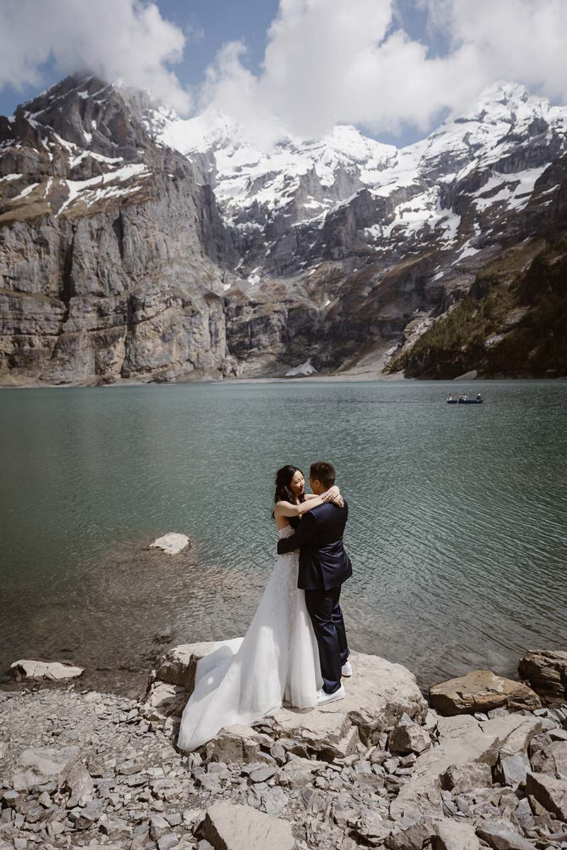 A bride and groom embrace on a rocky shore beside a lake with snow-capped mountains in the background—a stunning scene perfect for your Elopement Page.