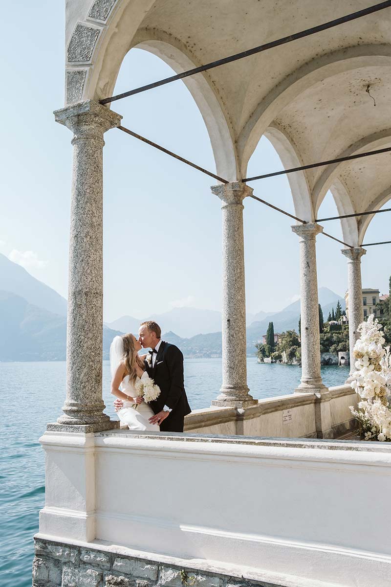 A bride and groom share a kiss under stone arches overlooking a lake, mountains, and lush greenery—a picture-perfect moment for your Elopement Page.