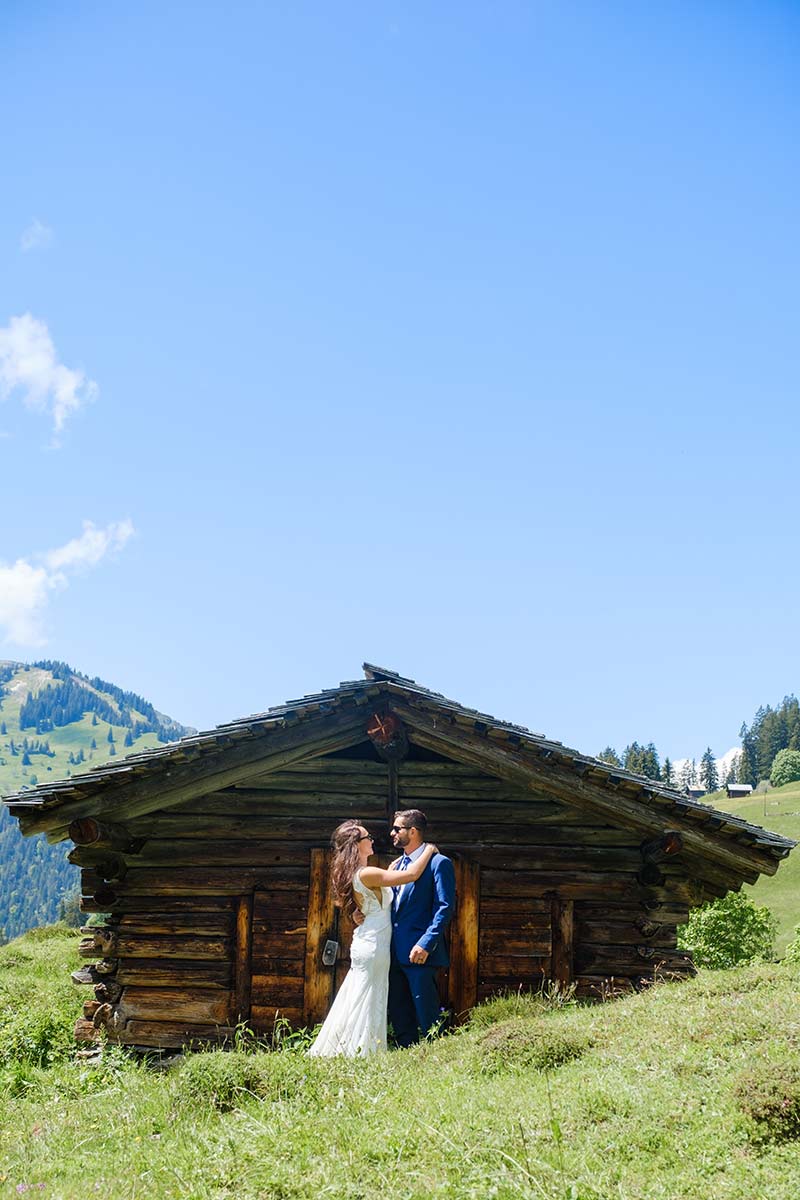 A bride and groom stand facing each other in front of a rustic Swiss cabin on a grassy hillside under a clear blue sky - the perfect setting for an intimate elopement.
