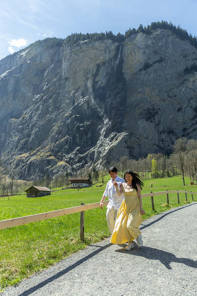 A man and woman walk on a gravel path through a grassy field in Wengen, with a rocky mountain and waterfall behind them—a perfect setting for proposal photography under the clear sky.