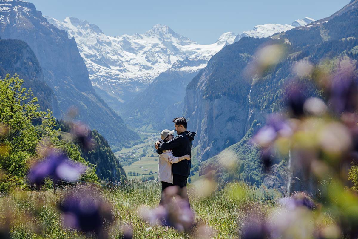 A couple embraces in a grassy field overlooking the stunning Lauterbrunnen Valley, with mountains and a valley in the background, beautifully framed by out-of-focus flowers—a perfect setting for proposal photography.