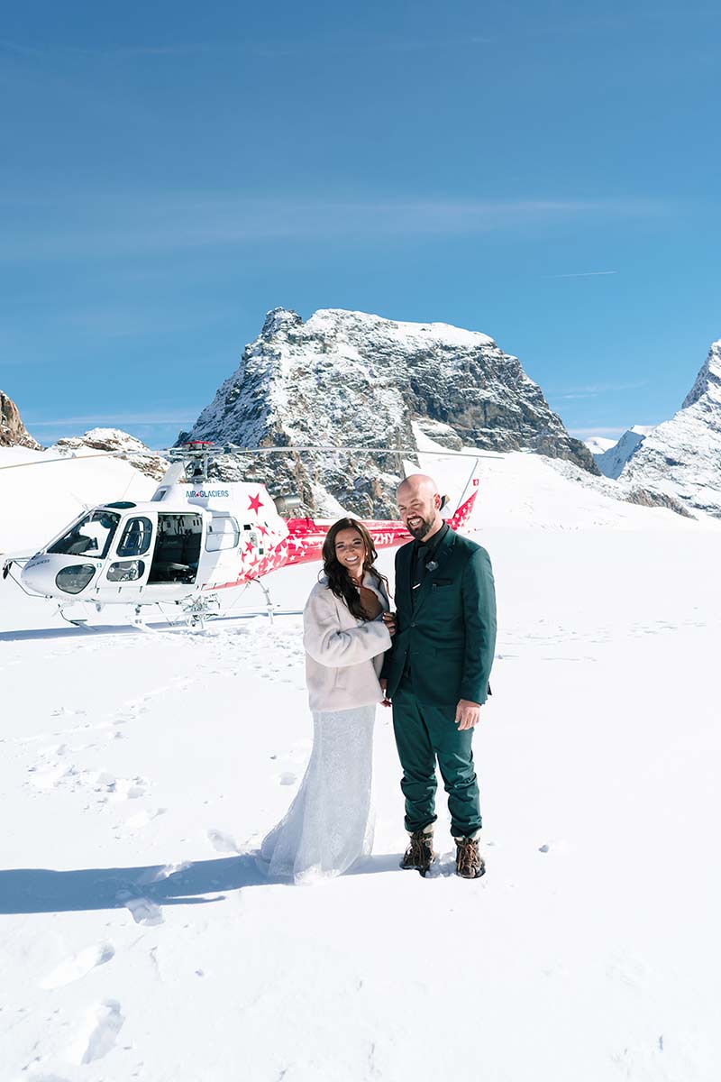 A couple dressed in formal attire stands on a snowy landscape in front of a helicopter, with snow-covered mountains in the background—a breathtaking way to elope in Switzerland.