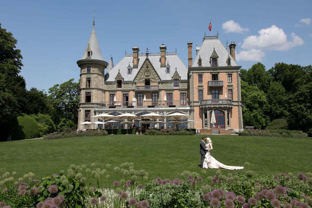 A bride and groom stand on the lawn in front of a large, ornate castle-like mansion with outdoor seating and landscaped gardens, making it the perfect spot to elope in Switzerland.