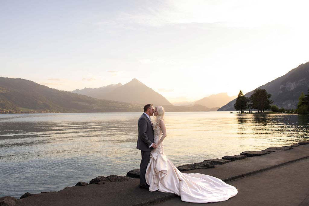 A bride and groom stand on a lakeside path at sunset, embracing and kissing with mountains and calm water in the background—capturing the magic of elope in Switzerland.
