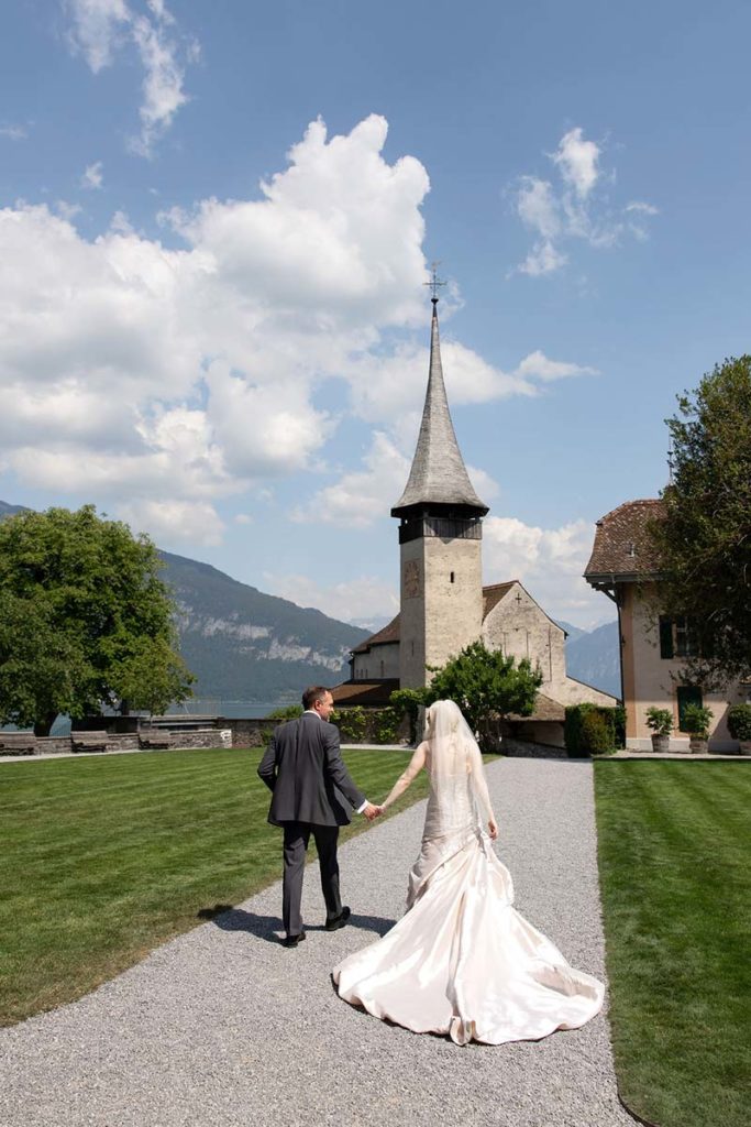 A bride and groom walk hand in hand on a gravel path toward a historic church, surrounded by green lawns and mountains under a partly cloudy sky—the perfect scene to elope in Switzerland.