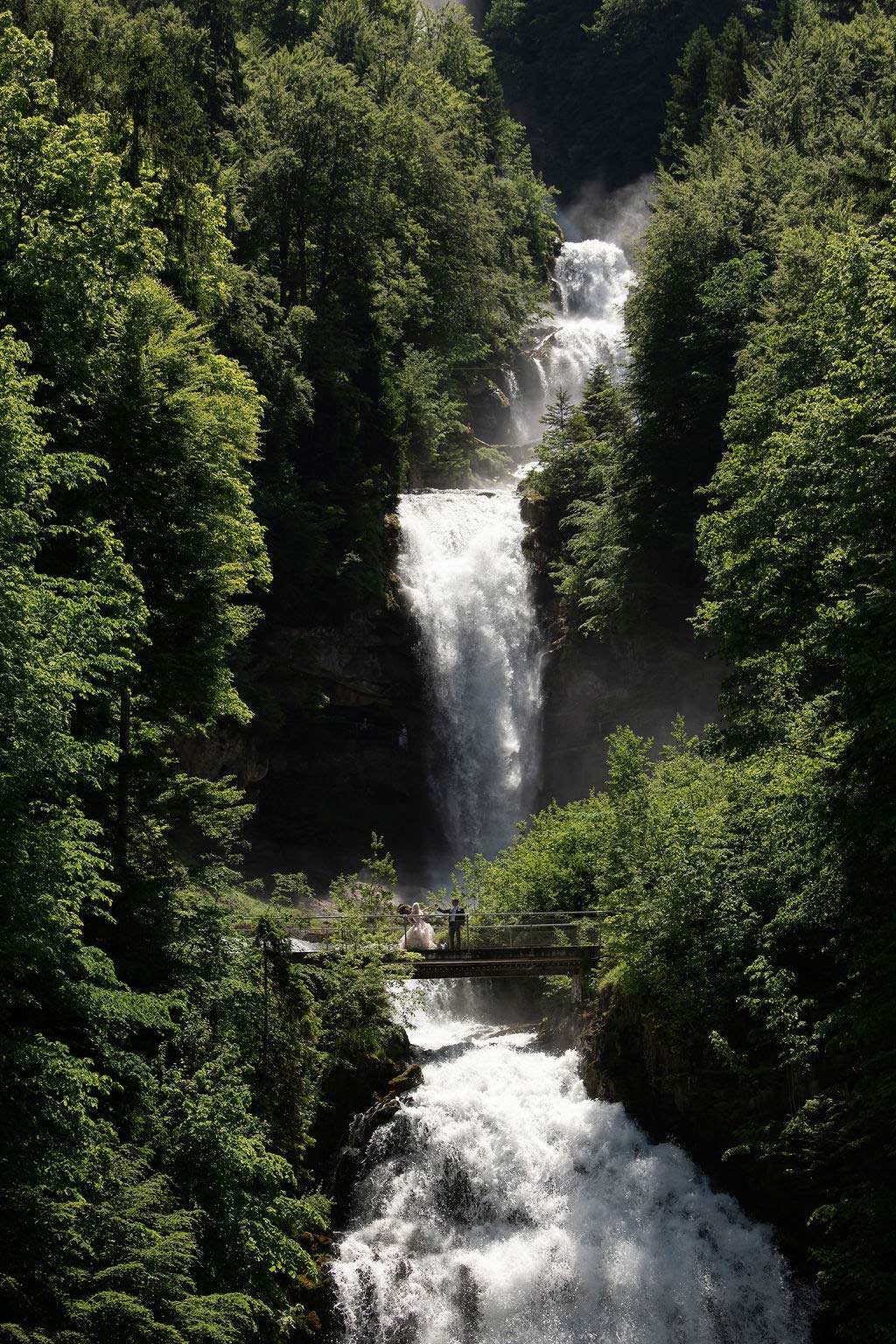 A tall, multi-tiered waterfall flows through dense green forest with a small bridge and people standing at its base—an enchanting spot for those dreaming of a Switzerland elopement.