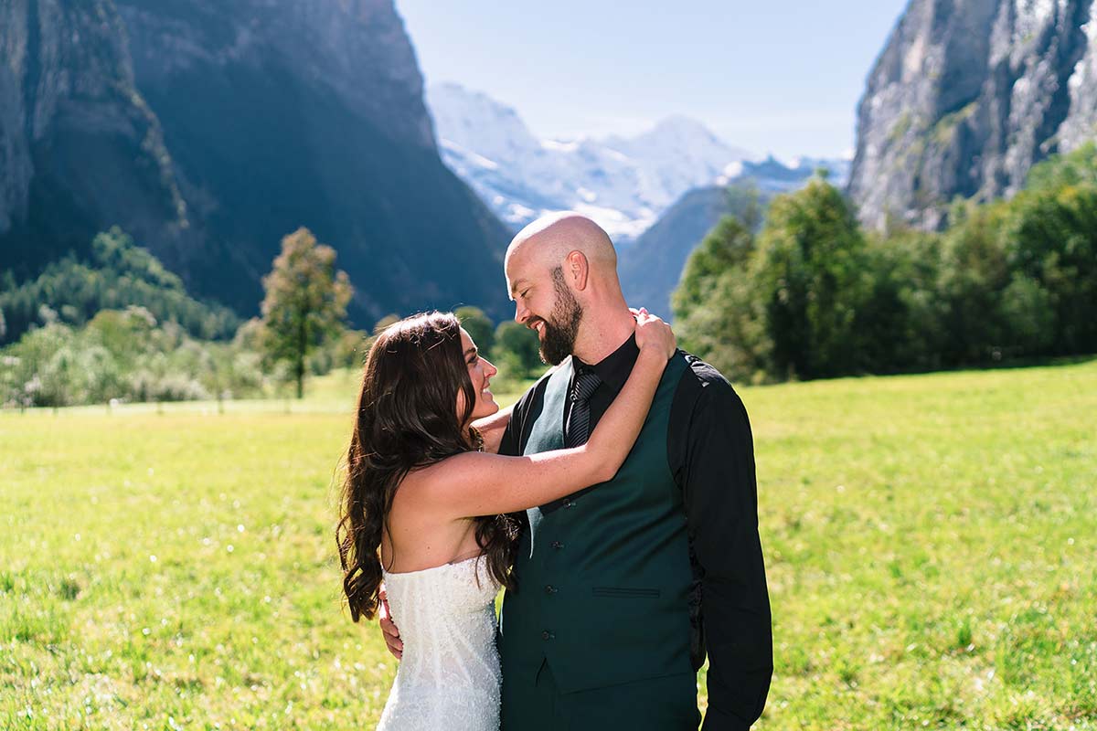 A couple dressed in wedding attire stands in a grassy field, embracing and smiling at each other, with mountains and trees framing their perfect moment—an ideal way to elope in Switzerland.