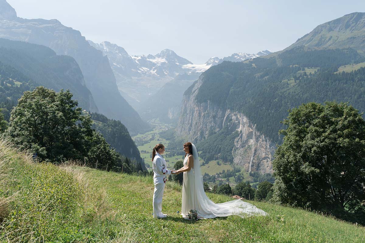 Two people stand facing each other in wedding attire on a grassy hill, ready to elope in Switzerland, with mountains and a valley in the background under a clear sky.