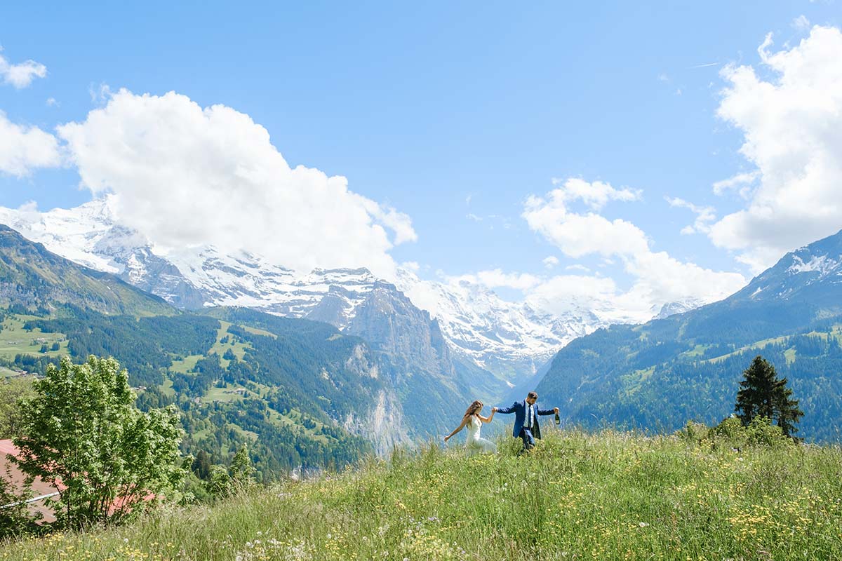 Two people walk hand in hand through a grassy meadow, with mountains and snow-capped peaks in the background - an idyllic scene to elope in Switzerland under a blue sky with scattered clouds.