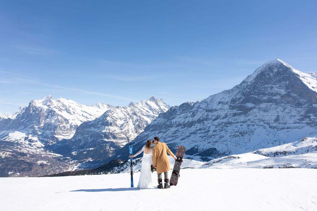 A couple in winter clothing stands on a snowy mountain slope with a snowboard, dreaming to elope in Switzerland as they gaze at distant snow-covered peaks under a clear blue sky.