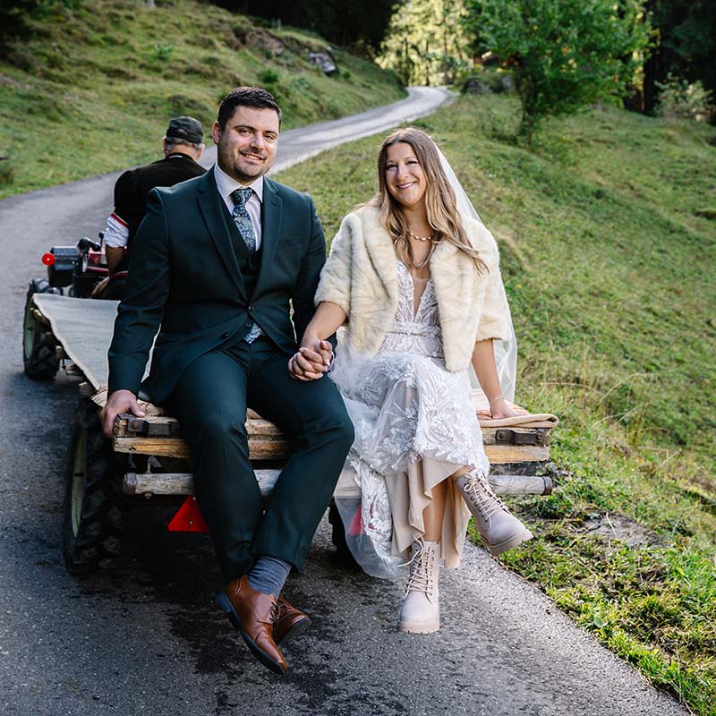 A bride and groom sit holding hands on a wooden cart towed by a small tractor along a rural road, surrounded by greenery - capturing the charm of elope in Switzerland.
