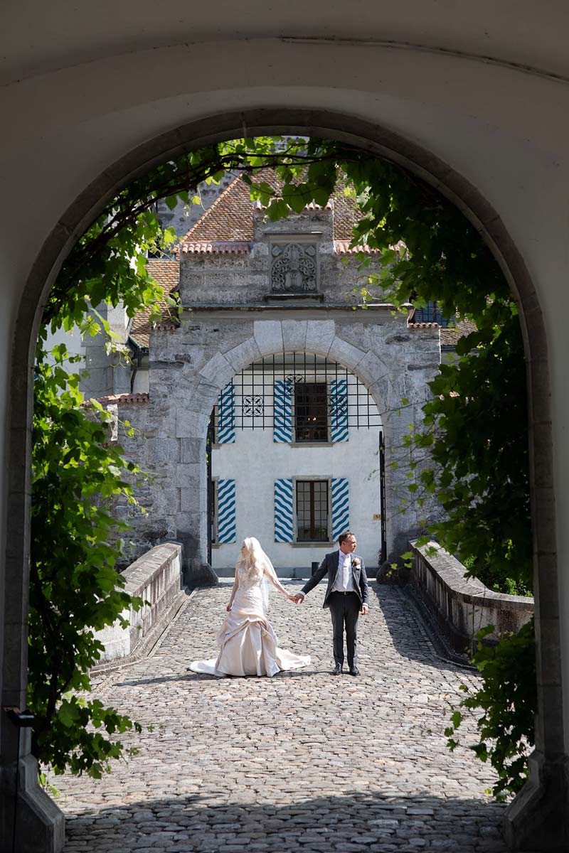 A bride and groom hold hands while walking across a cobblestone path toward a stone archway at a historic building, capturing the romance of an elope in Switzerland.