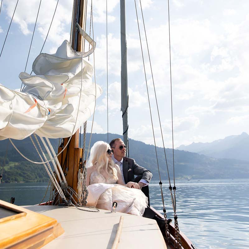 A bride and groom sit together on a sailboat, calm water and mountains in the background under a partly cloudy sky, capturing the romance of eloping in Switzerland.