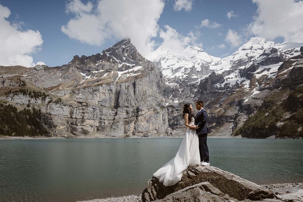A bride and groom stand on a rock beside a lake, surrounded by mountains with snowy peaks under a partly cloudy sky—an idyllic scene perfect for intimate elopement packages.