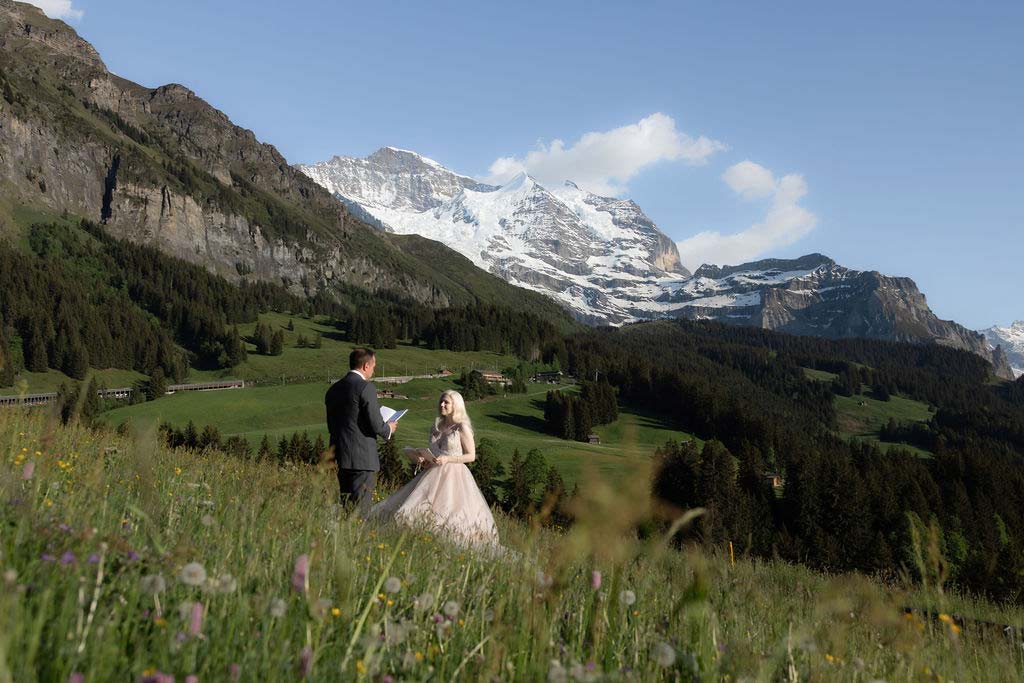 A couple stands in a mountain meadow, with the man reading from a paper, surrounded by wildflowers and snow-capped peaks—capturing the magic that elopement packages can offer in breathtaking natural settings.