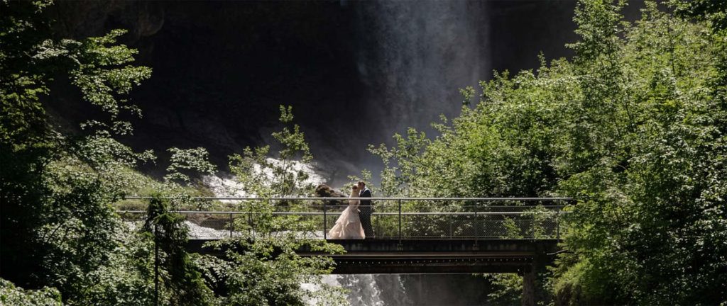 A couple stands on a narrow bridge surrounded by lush greenery with a waterfall in the background, reflecting the romance and adventure found in our elopement packages.