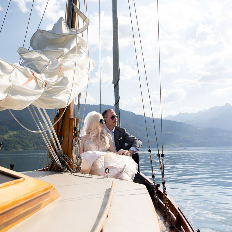 A couple dressed formally sits together on a sailboat, with mountains and water in the background under a partly cloudy sky—an intimate moment that captures the essence of elopement packages.