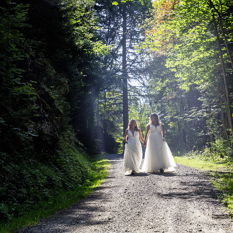 Two women in white dresses walk hand-in-hand along a sunlit, tree-lined gravel path in a forest, embodying the intimacy and beauty celebrated in our elopement packages.