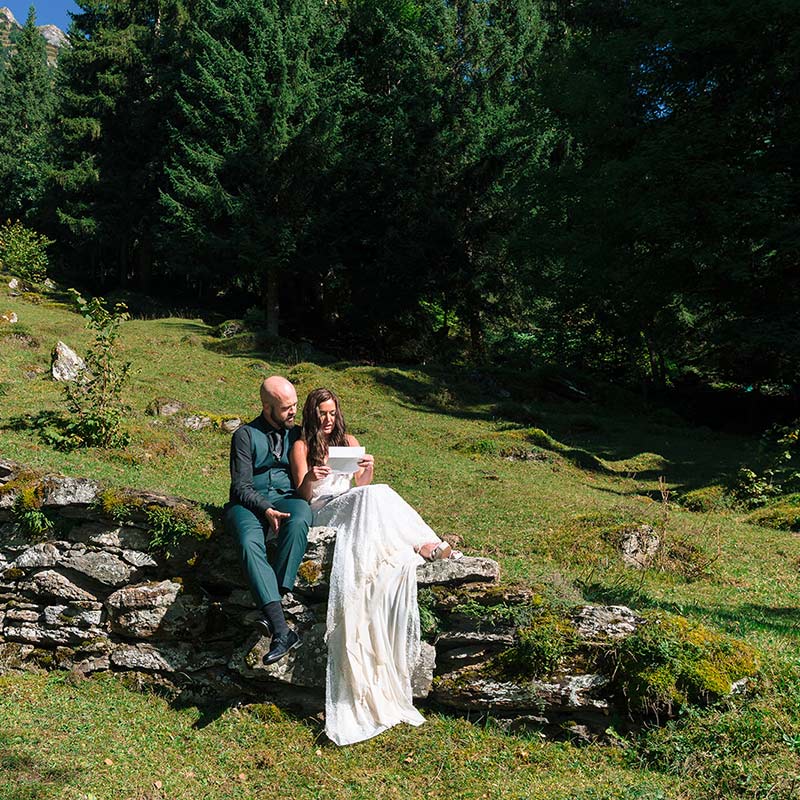 A couple dressed in formal attire sits on a stone wall in a grassy, wooded area, sharing an intimate elopement moment as the woman reads from a piece of paper.