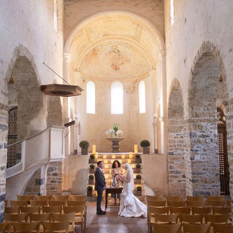 A bride and groom stand before an officiant in a stone-walled church with arched windows, wooden chairs, and decorative greenery—an intimate elopement setting perfect for exploring exclusive elopement packages and transparent pricing.