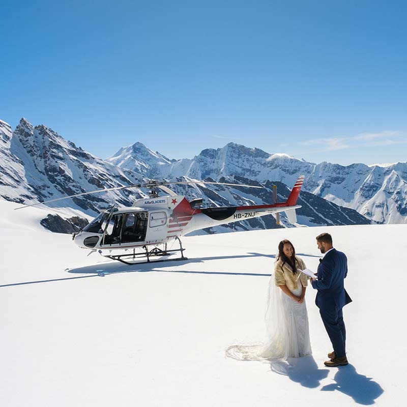A couple stands on a snowy mountain with a helicopter behind them, surrounded by snow-covered peaks under a clear blue sky—an unforgettable elopement adventure.