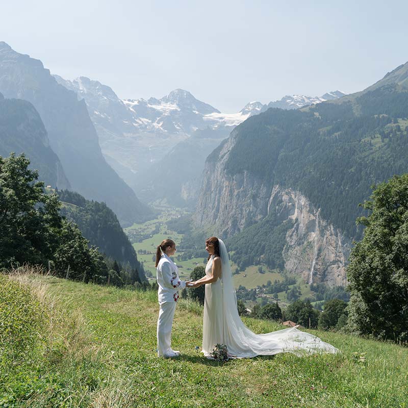 Two people in wedding attire stand facing each other on a grassy hillside, with mountains and a valley in the background, creating the perfect scene for couples exploring elopement packages or considering elopement cost for an intimate celebration.