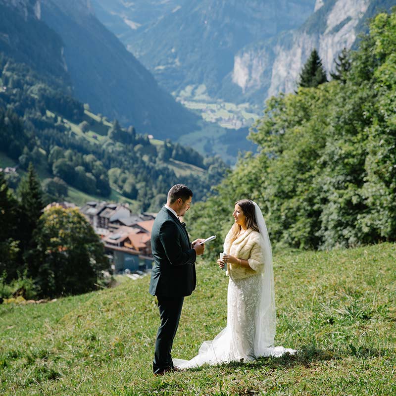 A bride and groom stand facing each other on a grassy hillside, exchanging vows, with a scenic mountain valley and village in the background—an intimate moment made possible by tailored elopement packages.