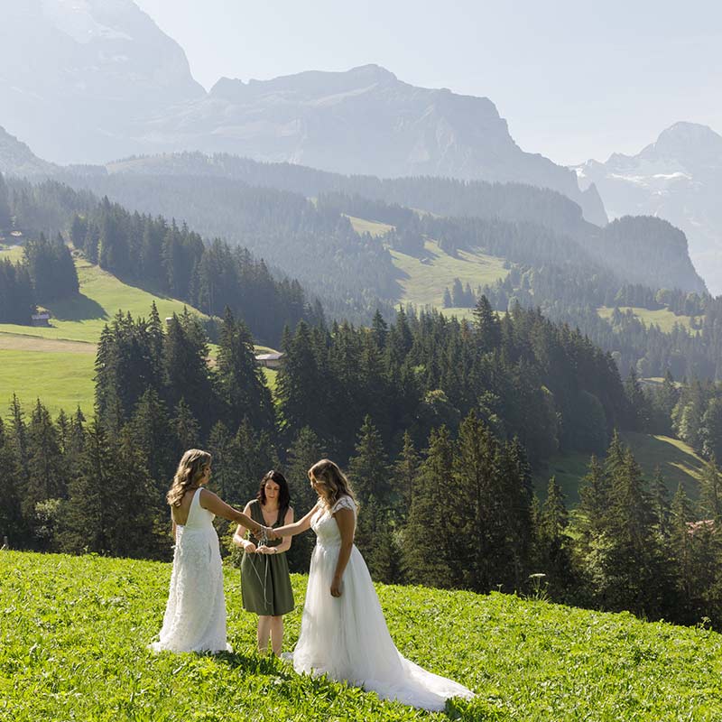 Two women in wedding dresses hold hands with an officiant in a green field, surrounded by mountains and trees—a beautiful moment that captures the romance of elopement packages and the freedom to focus on love over elopement cost.