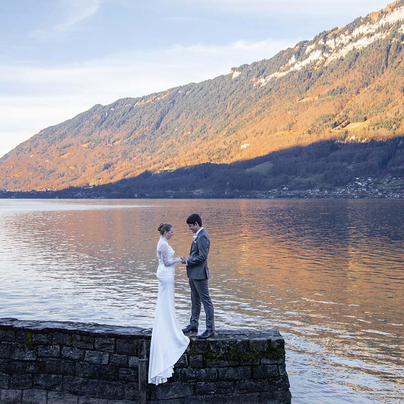 A bride and groom stand on a stone ledge by a lake with mountains in the background, holding hands and facing each other—an intimate moment that perfectly captures the magic of elopement packages.
