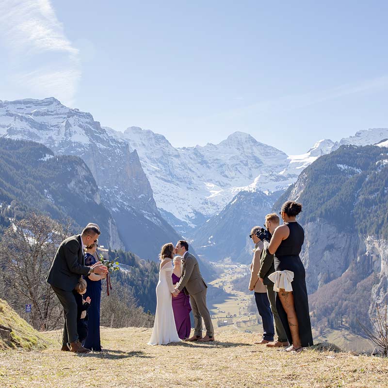 A couple kisses at an outdoor wedding ceremony in a mountain valley, surrounded by guests and photographers, with snow-capped peaks in the background—an unforgettable moment made possible by custom elopement packages.