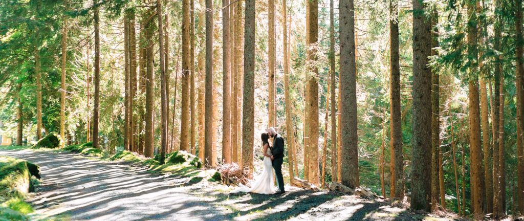 A couple in wedding attire stands close together among tall trees beside a sunlit forest path, capturing the magic and intimacy that make elopement packages such a wonderful choice for couples seeking a unique celebration.