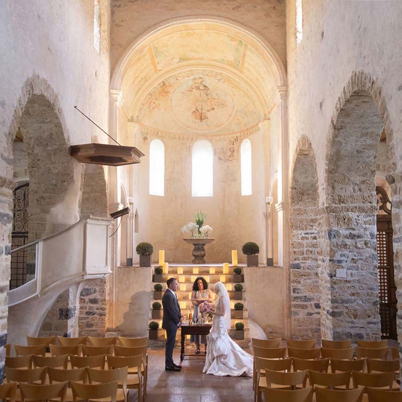 A couple stands before an officiant in a historic stone church with arched ceilings and wooden chairs, exchanging vows during an intimate Swiss elopement ceremony.