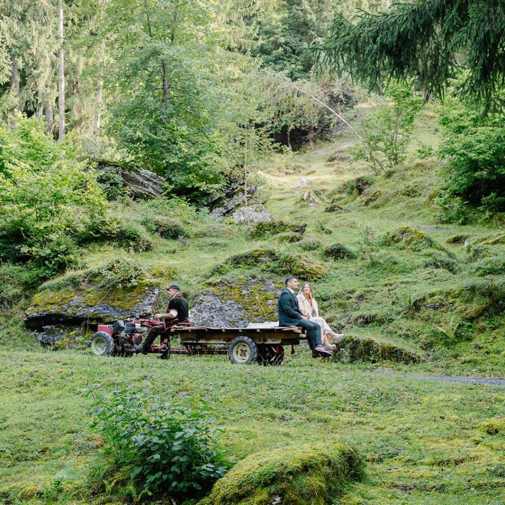 Two people sit on a wooden cart attached to a small tractor, surrounded by lush green forest and mossy rocks—a perfect setting for an intimate elopement adventure.