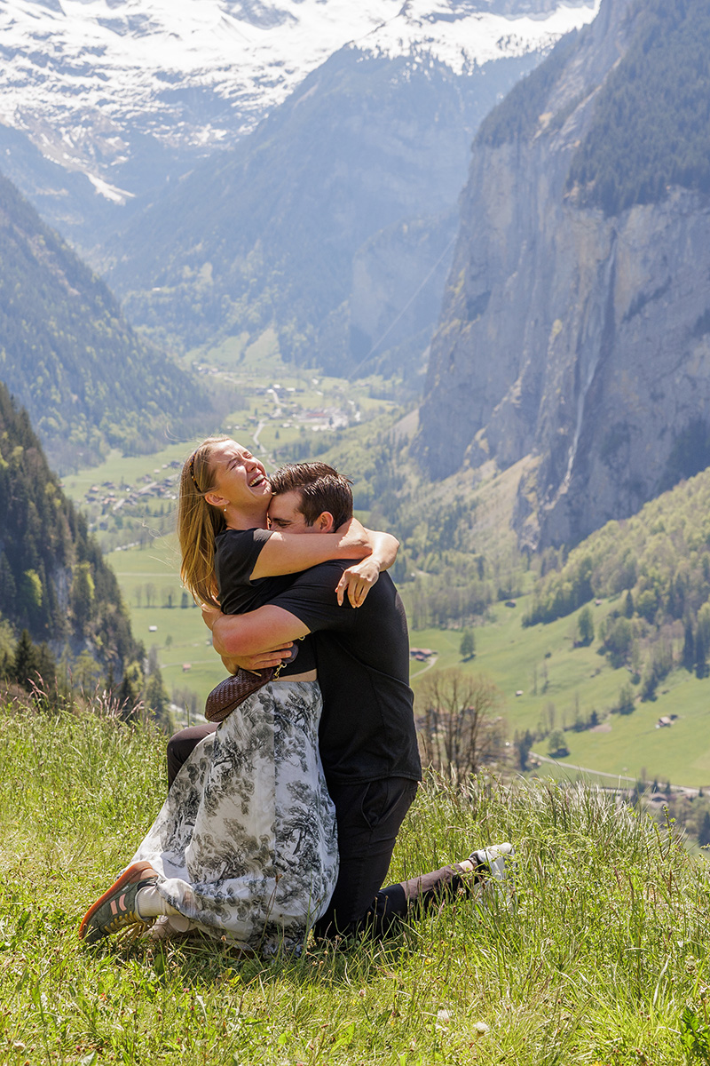 A couple embraces joyfully on a grassy hillside with a scenic valley, forest, and snow-capped mountains in the background—capturing a moment as memorable and full of promise as creating a New Draft on the Proposals Page.