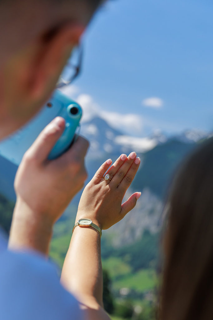 A person takes a photo of another person's hand wearing an engagement ring, with mountains and blue sky in the background, capturing a priceless memory that makes any proposal cost or plans worth it.