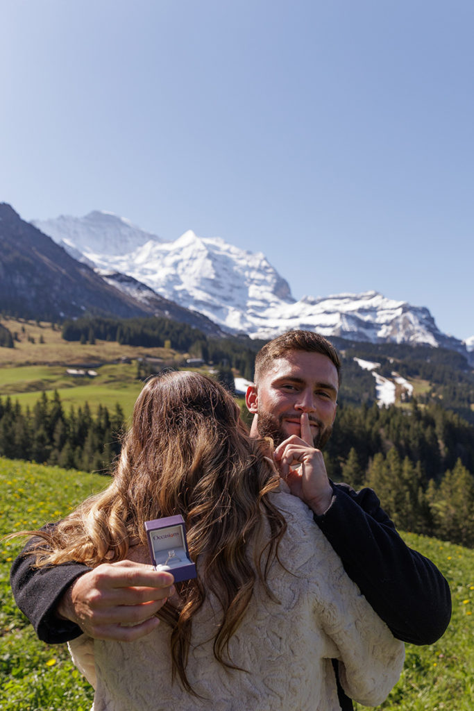 A man hugs a woman outdoors, holding an engagement ring box and signaling to be quiet, with snow-capped mountains in the background—capturing a moment as breathtaking as discovering the perfect proposal pricing page for your special day.