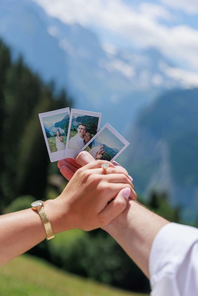 Two hands hold each other, with one hand also holding three instant photos against a blurred mountain and forest landscape in the background—capturing a moment as memorable and meaningful as choosing the perfect proposal.