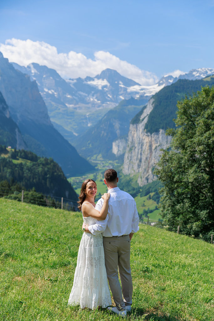 A couple stands on a grassy hill overlooking a valley with mountains and cliffs in the background under a blue sky, sharing a moment as memorable as the first look at a new draft on the proposals page.