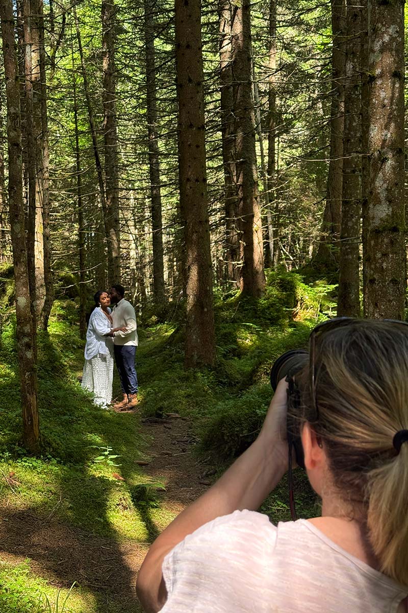 A photographer takes a picture of a couple standing together in a sunlit forest surrounded by tall trees and green moss, capturing the kind of natural beauty perfect for featuring on your Proposals Page or in New Draft proposals.