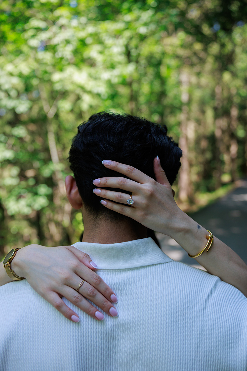 A person with dark hair is seen from behind, embraced by another whose hands, wearing gold rings and a bracelet, are resting on their neck and shoulder. Trees and a path are in the background, suggesting a quiet moment away from the bustle of drafting new proposals or refining ideas for the Proposals Page.