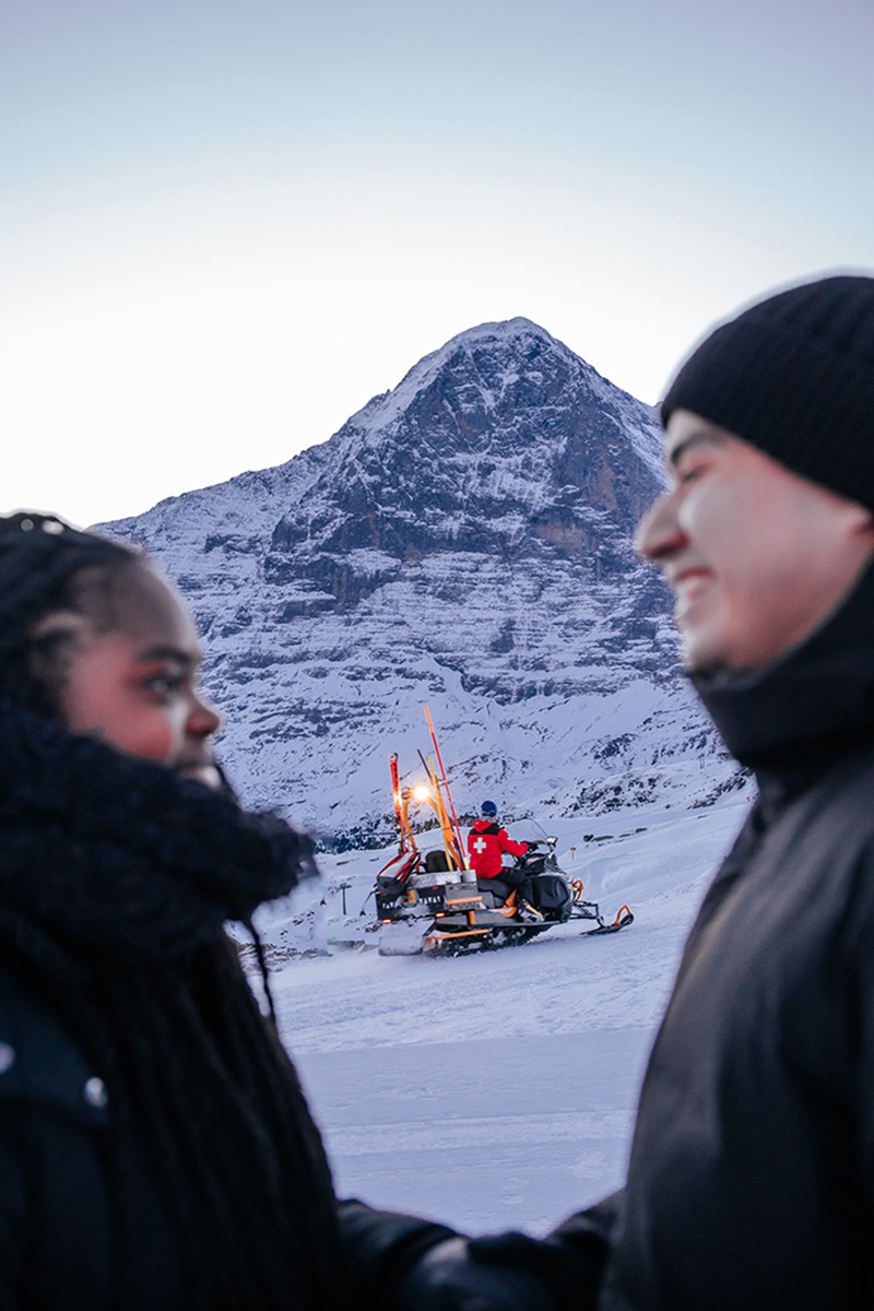 Two people in winter clothing stand in focus in the foreground, resembling collaborators reviewing a New Draft, while a person on a snowmobile is seen traversing the snowy mountain landscape in the background.