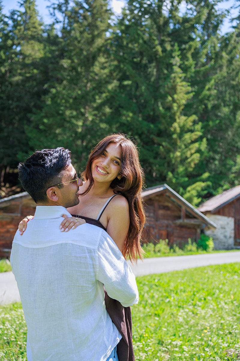 A man in a white shirt lifts a smiling woman in a sundress outdoors, with wooden cabins and tall green trees in the background, capturing a joyful moment reminiscent of saying 'yes' to the perfect proposal.