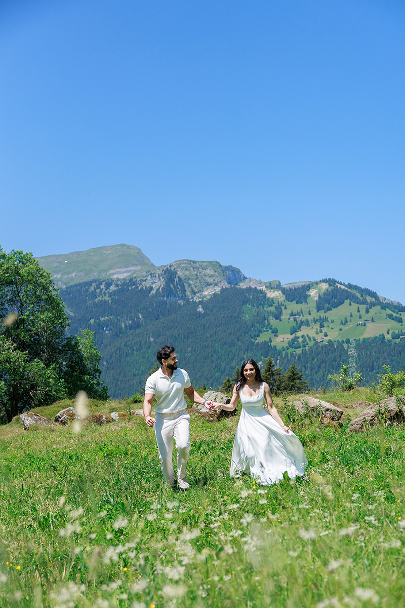 A couple in white outfits walks hand in hand through a green meadow with mountains and blue sky in the background, evoking the fresh possibilities of a New Draft on the Proposals Page.
