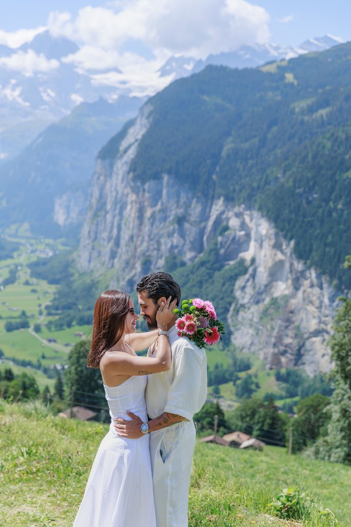 A couple in white stands embracing in a meadow with a bouquet of flowers, set against a backdrop of the lush green Lauterbrunnen Valley and snow-capped peaks in Switzerland—a breathtaking scene perfect for proposal photography.