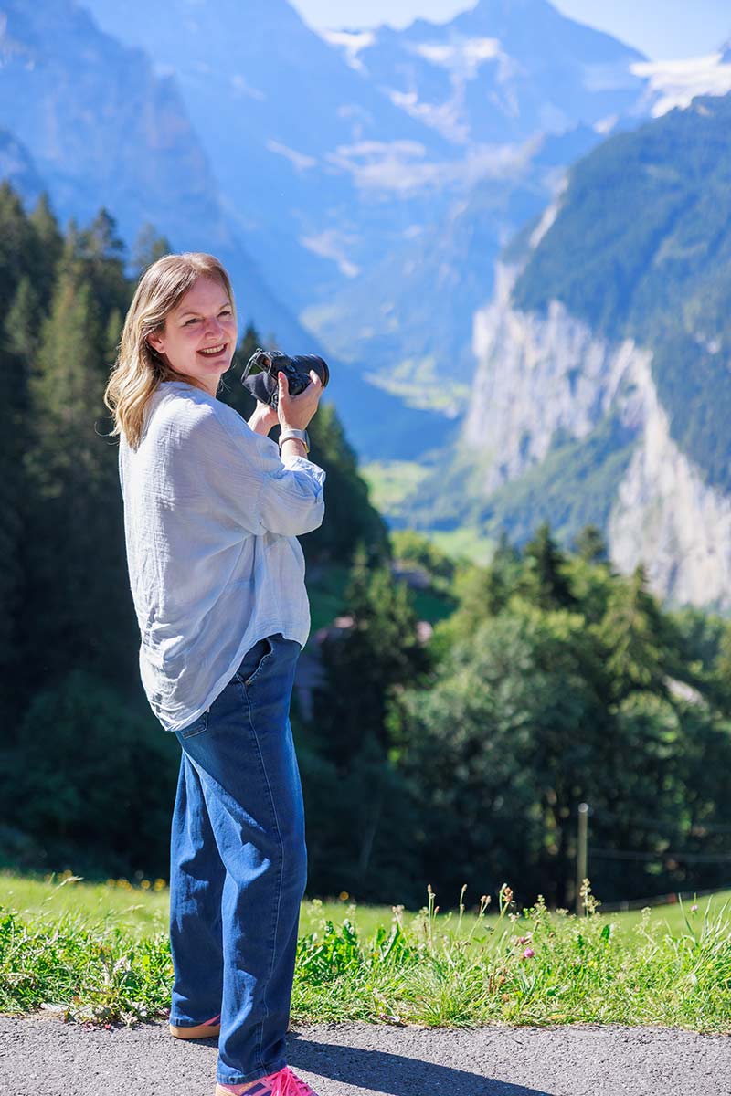 A woman holding a camera stands on a grassy path in Switzerland with a mountainous valley in the background.